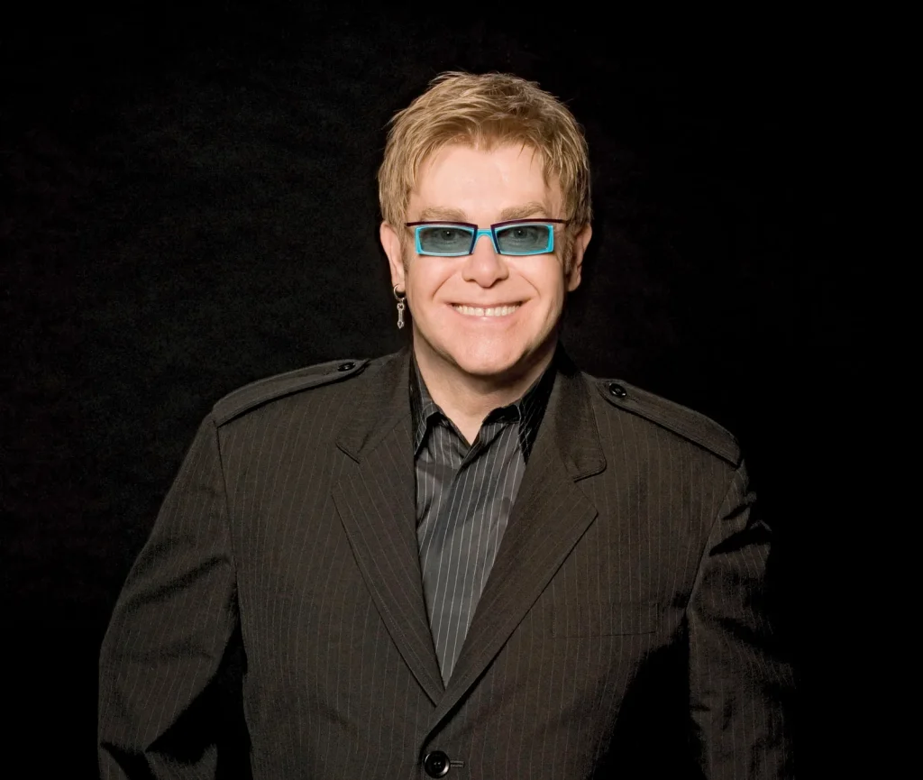 Smiling Elton John grey suit with matching lining shirt and blue spectacles - Famous people with lasik 