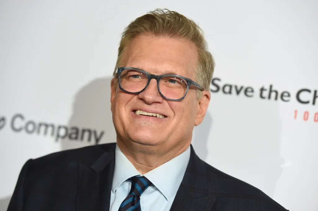 Smiling Drew Carey in black coat with sky blue shirt with matching tie and spectacles - Famous people with lasik 