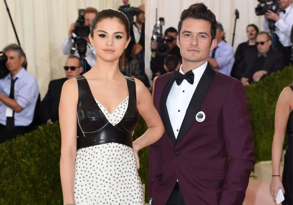 Selena Gomez in white polka dotted dress with black waste coat and Orlando Bloom in purple suit with white shirt posing for camera Selena Gomez in white polka dotted dress with black waste coat and Orlando Bloom in purple suit with white shirt posing for camera - selena gomez age