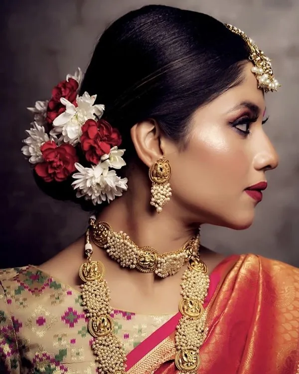 A girl in orange saree with multicolor blouse and heavy golden jewellery posing for camera and showing the side view of her Tight Bun Adorned with Artificial Flowers A girl in orange saree with multicolor blouse and heavy golden jewellery posing for camera and showing the side view of her Tight Bun Adorned with Artificial Flowers - hairstyles for Indian Wedding