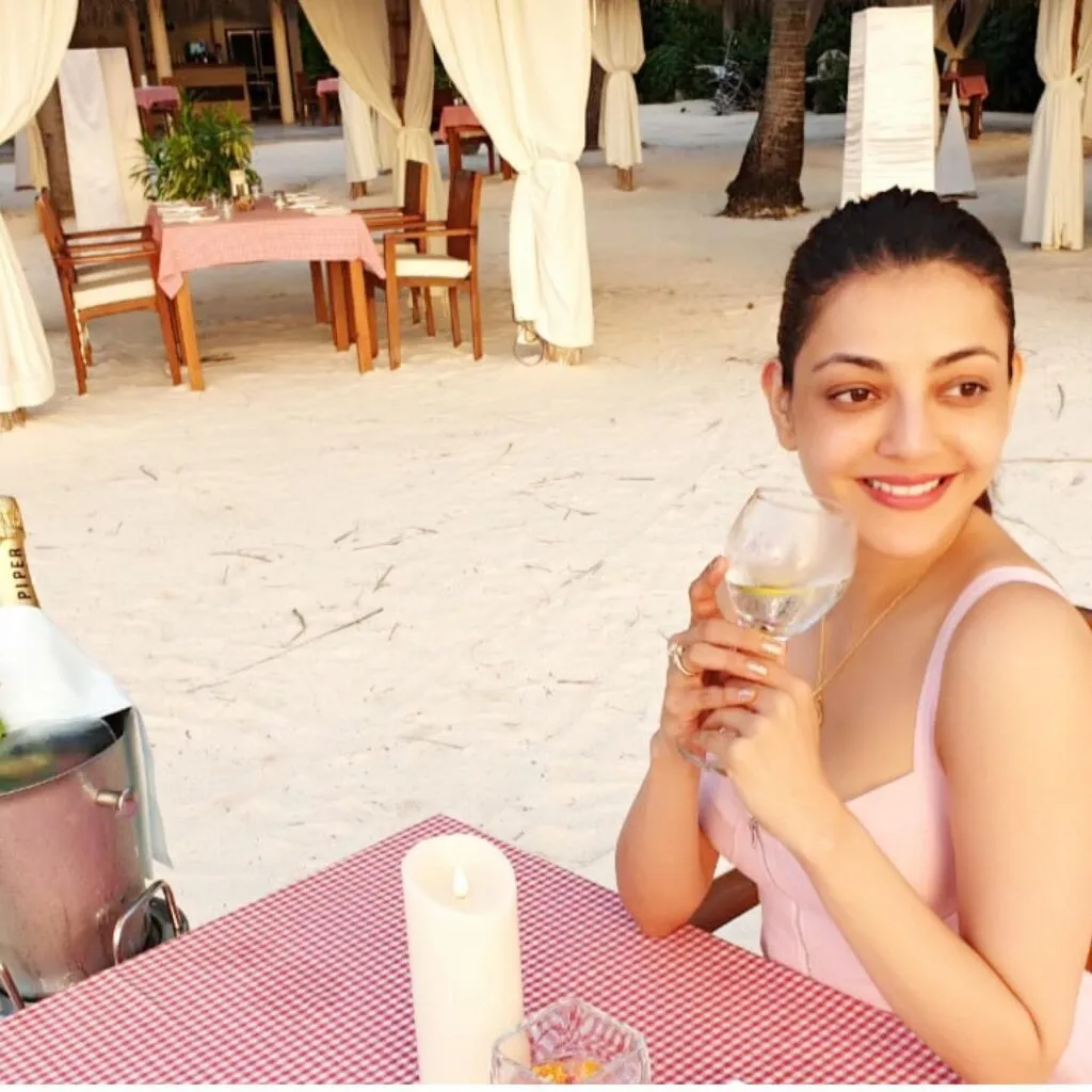 Smiling Kajal Agarwal sitting on a chair with a glass of drink in pink strappy dress Smiling Kajal Agarwal sitting on a chair with a glass of drink in pink strappy dress - south actress without makeup